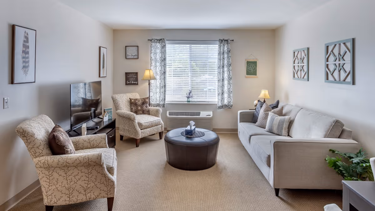 A cozy living room with a beige sofa on the right, two patterned armchairs on the left, a round dark ottoman in the center, and a flat-screen TV on a stand against the left wall. The room has a large window with patterned curtains, two table lamps, wall art, and a small plant on the right side.