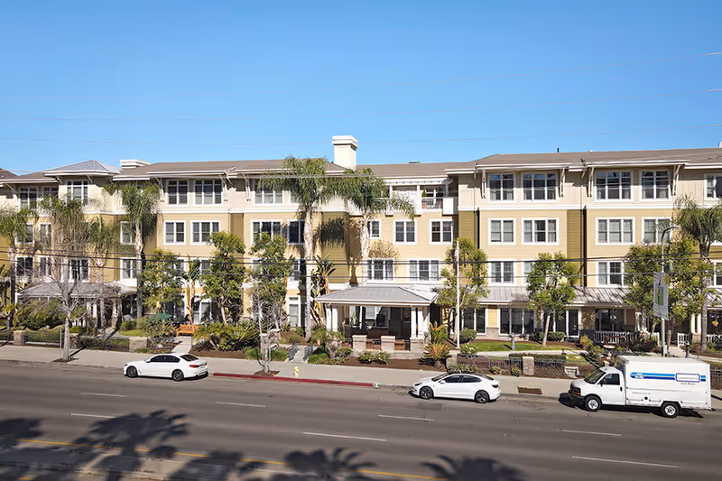 Front exterior of a multi-story senior living building with palm trees and parked cars along the street.