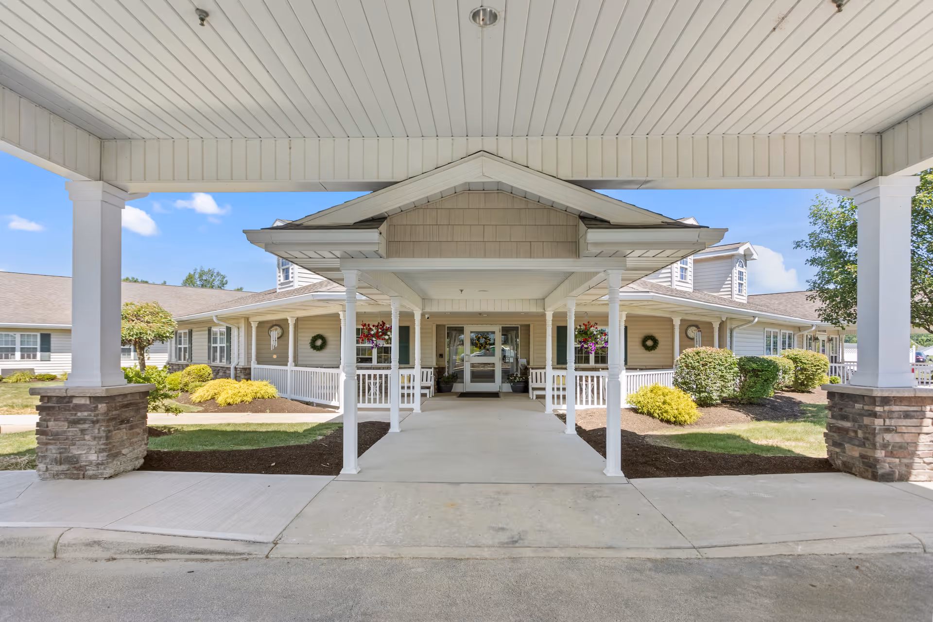 Covered entrance of a single-story nursing facility building with white pillars, stone bases, and a walkway leading to glass double doors. The building is surrounded by neatly trimmed bushes and flower pots hanging near the entrance under a clear blue sky.