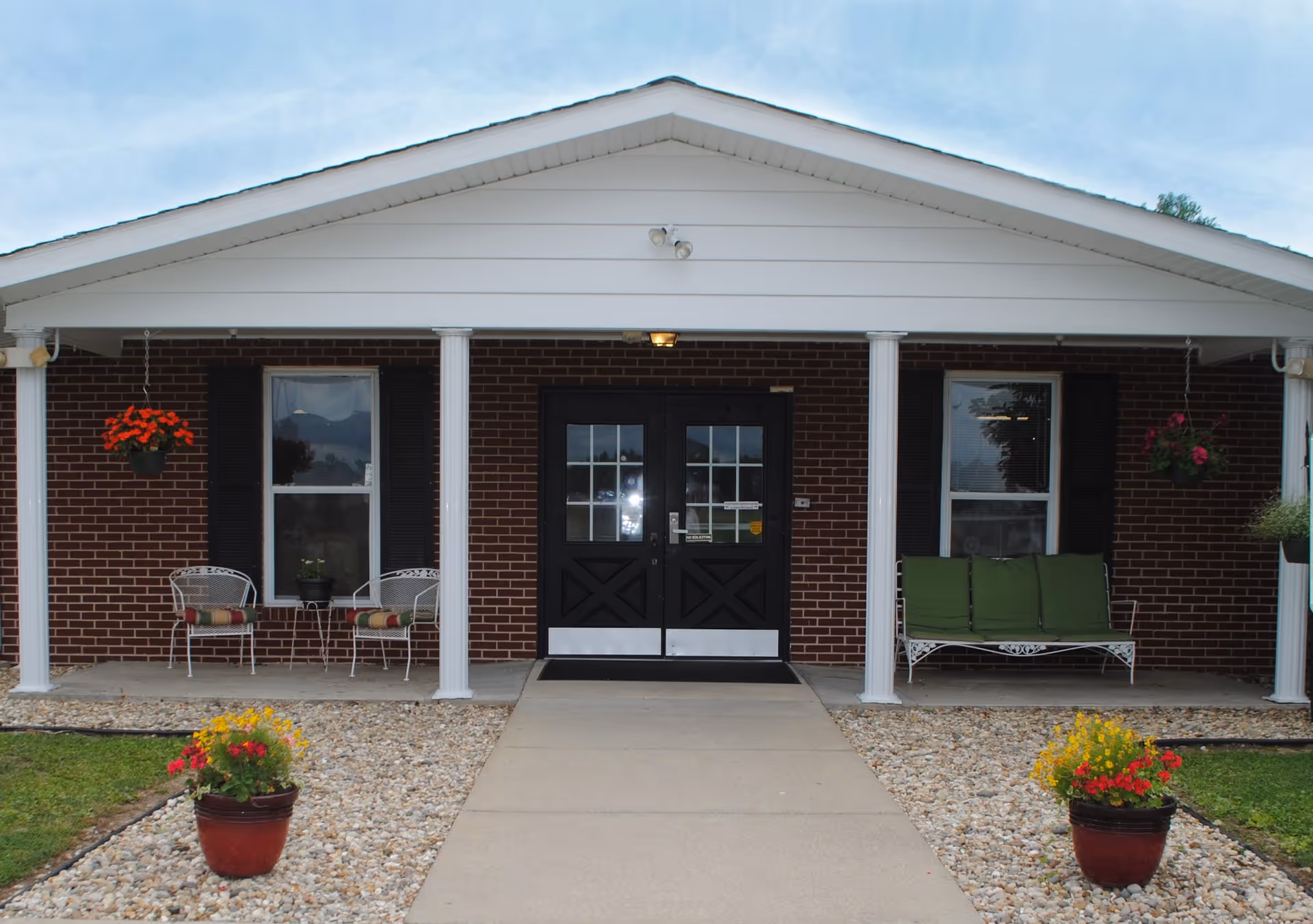 Front entrance of a brick senior living building with a covered porch, white columns, double black doors, seating and potted flowers.