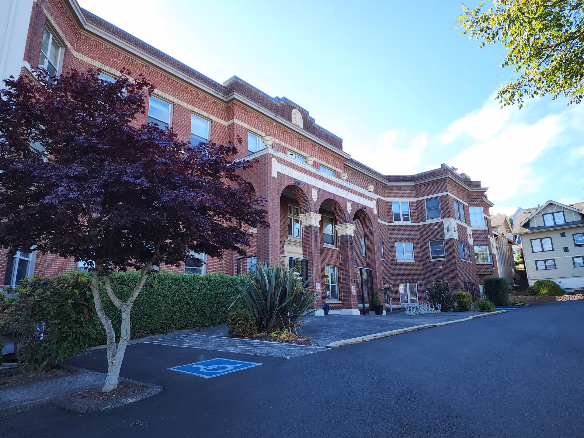 Exterior view of a large, three-story brick building with arched entrance and decorative architectural details. There is a tree with dark red leaves and some shrubs near the entrance. The sky is clear and blue, and there is a handicap parking space visible in the foreground.