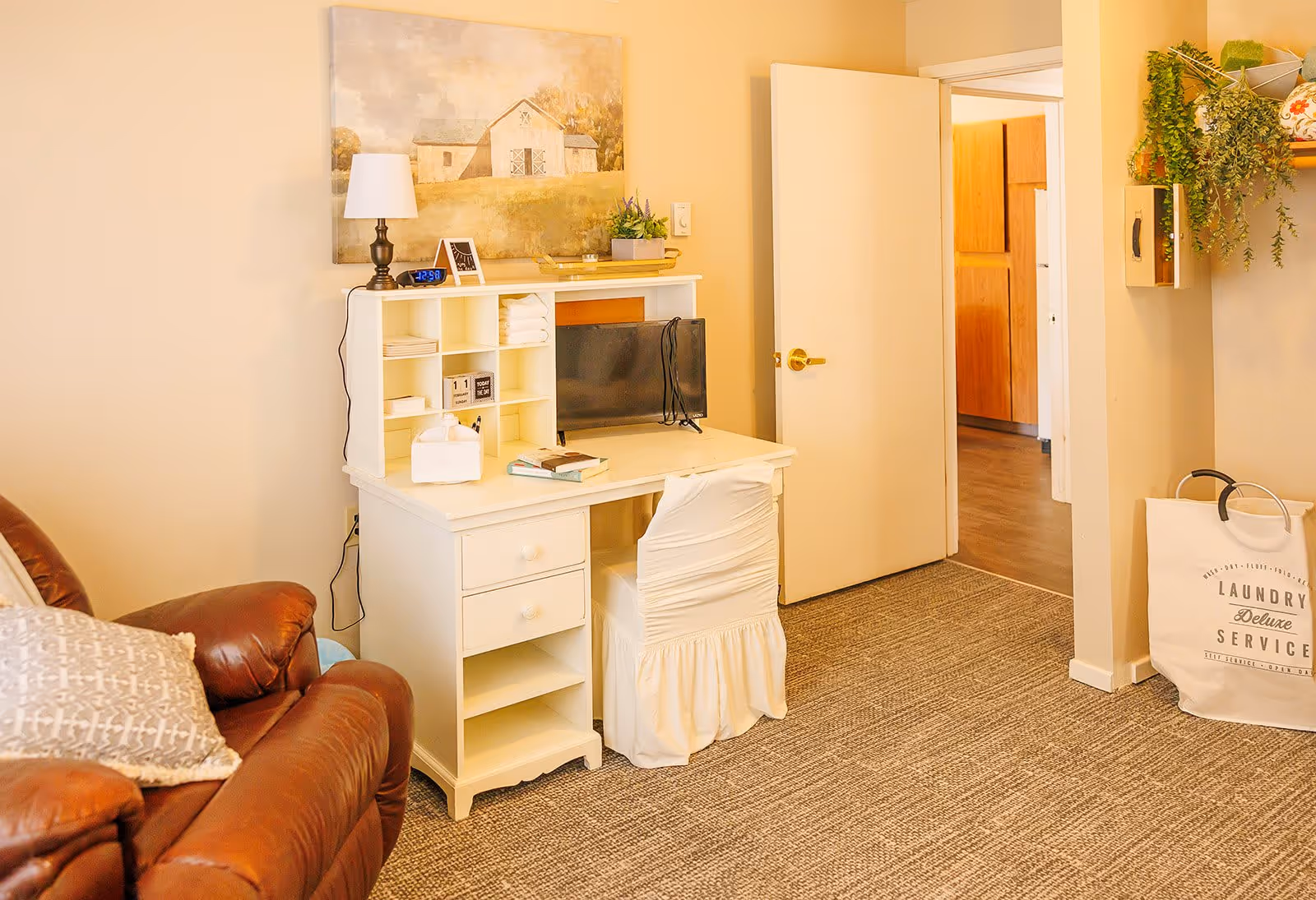 A cozy room with a brown leather armchair on the left and a white desk with a matching chair covered in a white slipcover. On the desk are a small TV, a lamp, a clock, books, and decorative items. A painting of a barn hangs on the wall above the desk. To the right, there is an open door leading to another room with wooden cabinets. A laundry bag labeled 'Laundry Deluxe Service' is on the floor near the wall with hanging greenery above it.