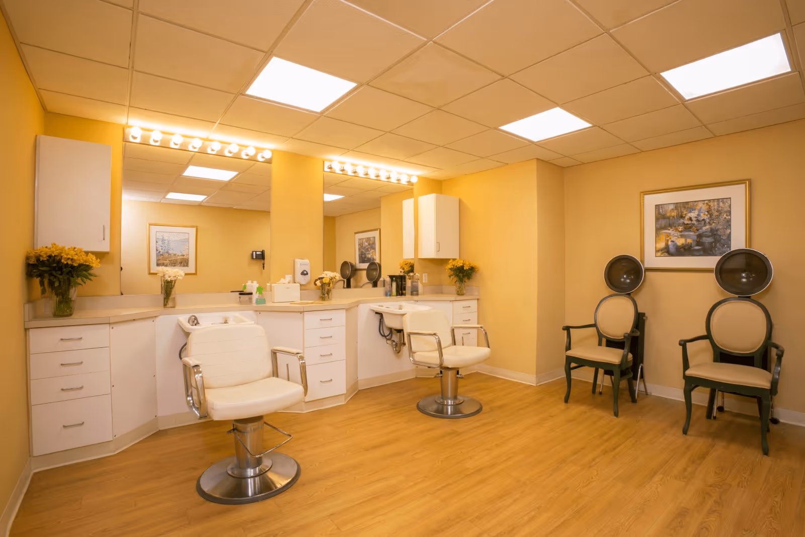 Interior of a hair salon area in a senior living facility with two white salon chairs in front of a counter with mirrors and bright vanity lights. There are two vintage hair dryers with chairs against a yellow wall, decorated with framed artwork and vases of flowers.