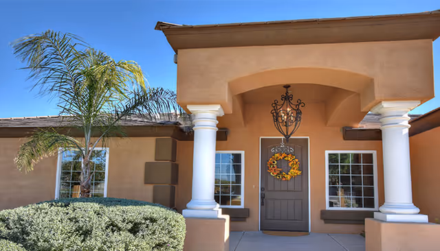 Front entrance of a stucco single-story building with white columns, a brown door adorned with a wreath, windows, and landscaping.