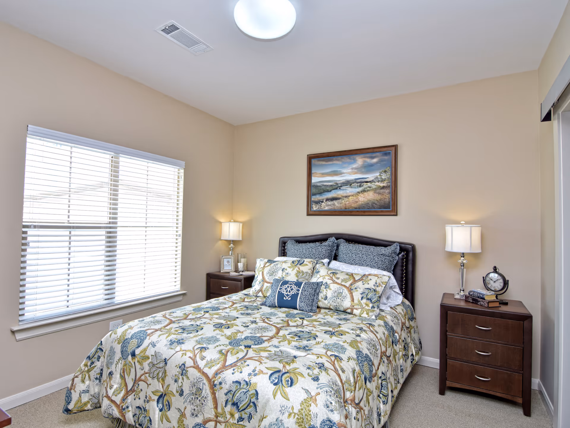 A bedroom with a bed covered in a floral patterned comforter and multiple pillows. There are two wooden nightstands on either side of the bed, each with a lamp. One nightstand has a clock and some books, while the other has a framed photo and a glass. A window with white blinds is on the left wall, and a framed landscape painting hangs above the bed.