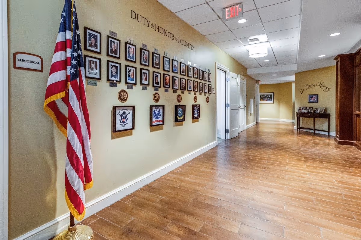 A hallway in a senior living facility with wooden flooring and beige walls. On the left wall, there is a display of framed photographs and plaques under the words 'DUTY * HONOR * COUNTRY' with an American flag standing beside it. At the far end of the hallway, there is a small table with framed pictures and the words 'In Loving Memory' on the wall above it. The ceiling has recessed lighting and a hanging light fixture, with an exit sign visible.