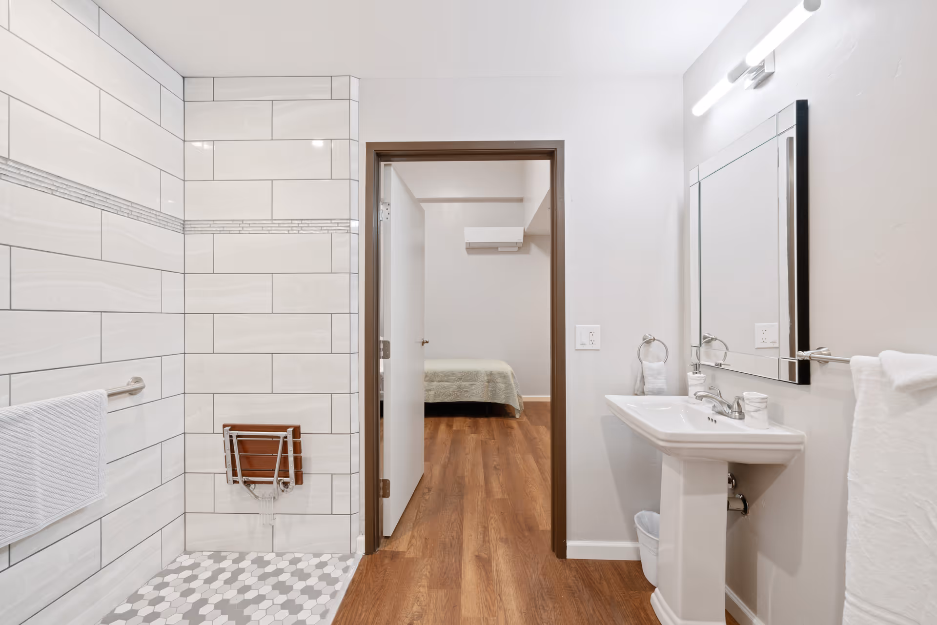A clean, modern bathroom with a walk-in tiled shower featuring a fold-down wooden seat and grab bar. The bathroom has a pedestal sink with a mirror above it, a towel ring with a hand towel, and a towel bar with a white towel. The floor is wood-style laminate, and through an open door, a bedroom with a bed and air conditioning unit is visible.