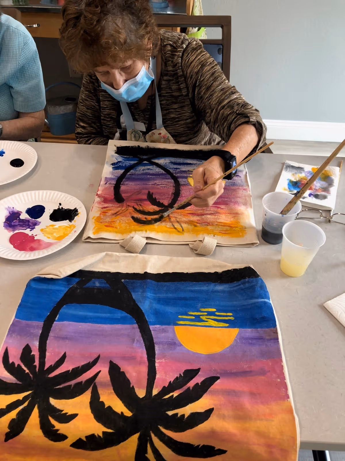 An elderly woman wearing a face mask and a brown patterned shirt is painting a colorful sunset scene with black palm trees on a canvas bag. There are paint palettes with various colors, brushes, and cups with water on the table.