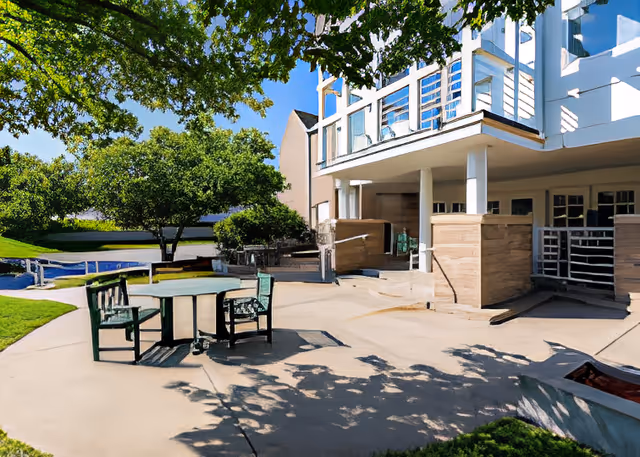 Outdoor patio area at Hickory Villa Assisted Living with a round table and four chairs on a concrete surface. The area is shaded by a large tree, and the building features a balcony with white railings and pillars. Green grass and additional trees are visible in the background under a clear blue sky.