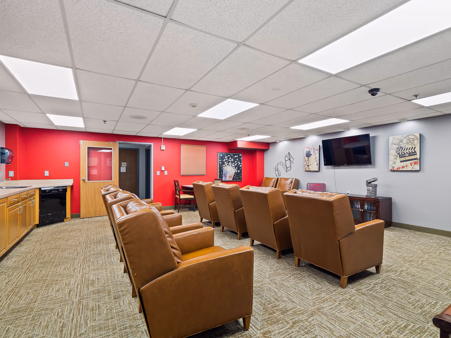 A cozy common room with two rows of brown leather armchairs facing a wall-mounted flat-screen TV. The room has a red accent wall with a wooden door and a small table with chairs. The opposite wall is gray with movie-themed decorations including film reels and popcorn artwork. There is a kitchenette area with wooden cabinets and a dishwasher on the left side.