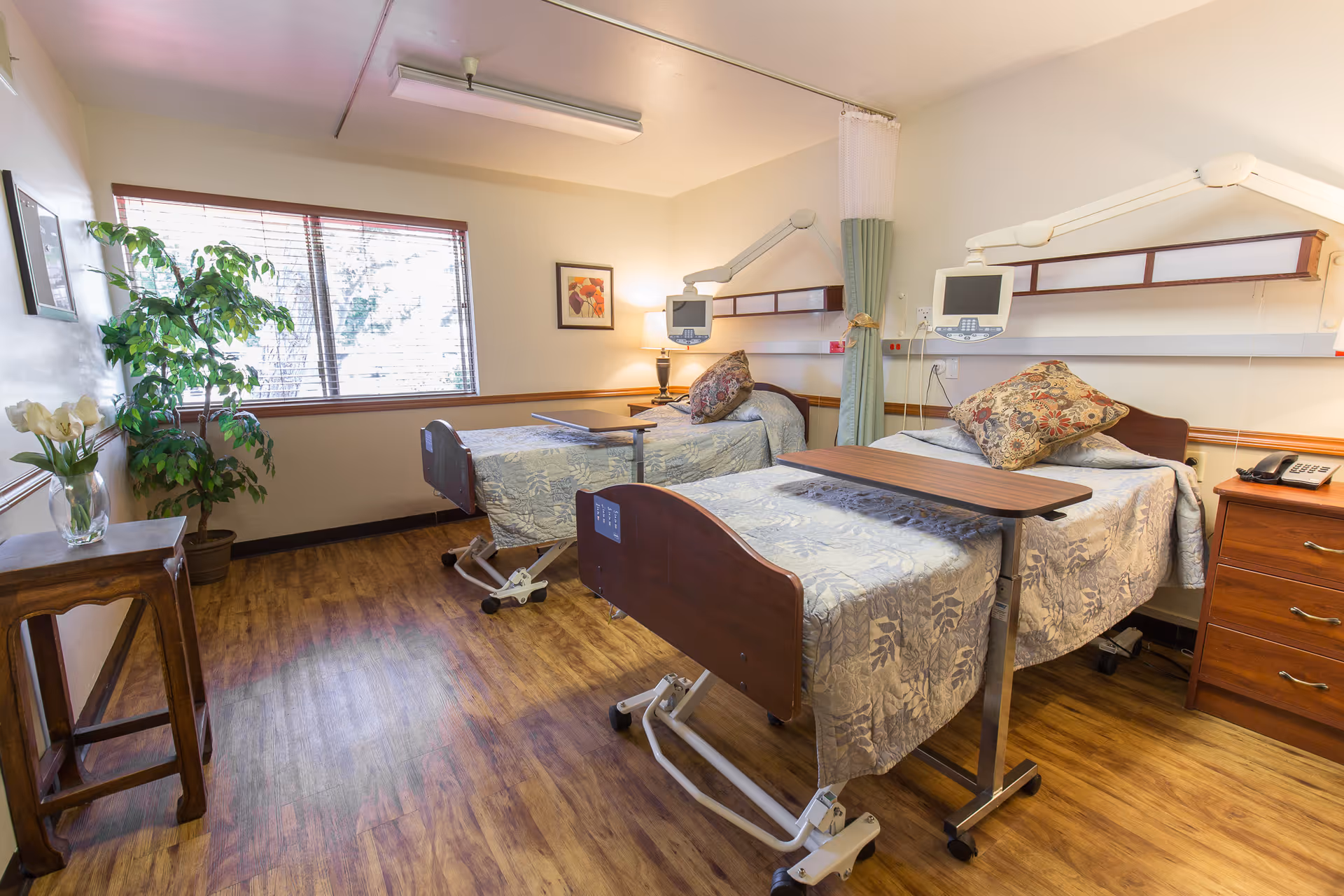 A bright and clean hospital-style room with two adjustable beds, each with a floral pillow and a light blue patterned bedspread. Each bed has an overbed table and medical monitoring equipment mounted on the wall above the headboards. The room has wooden flooring, a large window with blinds, a potted plant, a small wooden table with a vase of flowers, a nightstand with a telephone, and a framed picture on the wall.