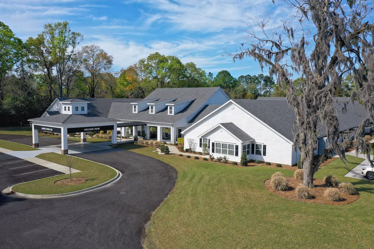 Front exterior of a single-story white senior living building with a covered entrance, curved driveway, and landscaped lawn.