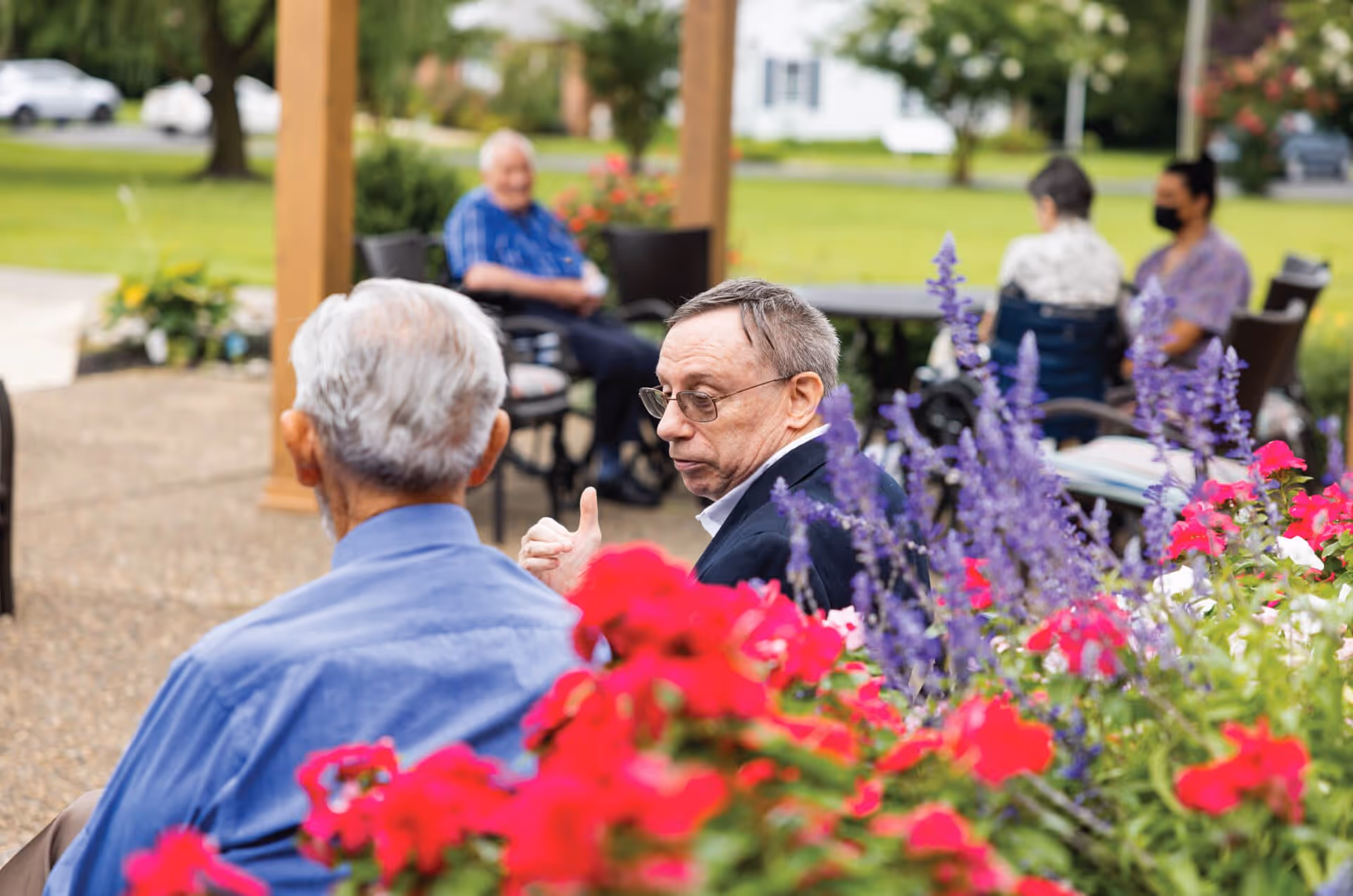 Several elderly individuals sitting outdoors in a garden area with colorful flowers in the foreground. Some people are seated in wheelchairs, and one person is wearing a face mask. The setting appears to be a peaceful outdoor space with greenery and a building in the background.