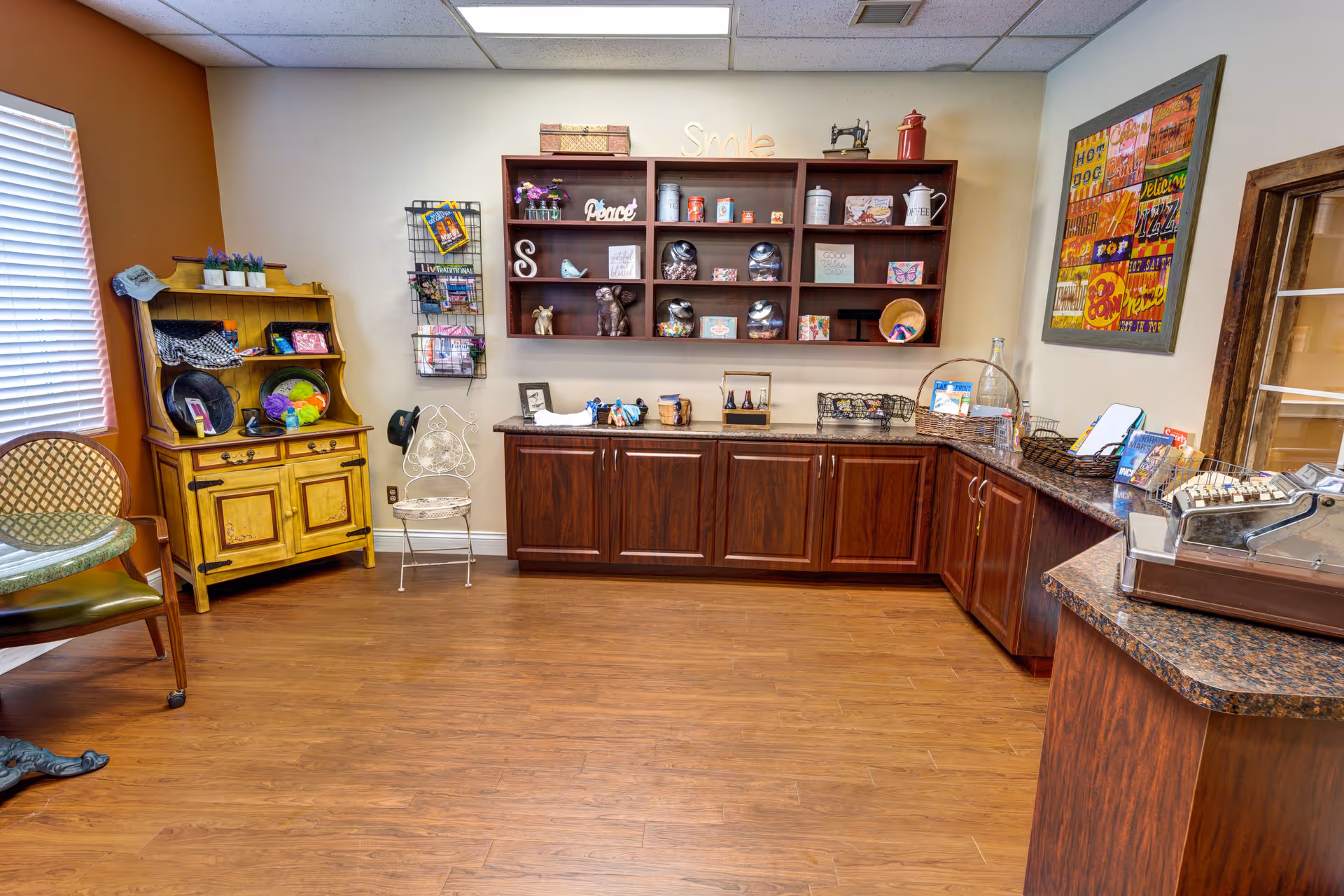Interior room with wooden flooring and cabinetry. There is a yellow wooden hutch with various items on it, a white decorative chair, and a wall-mounted shelf holding decorative objects and signs. A colorful framed artwork hangs on the wall above a granite countertop with baskets and a vintage cash register. A window with blinds is on the left side.