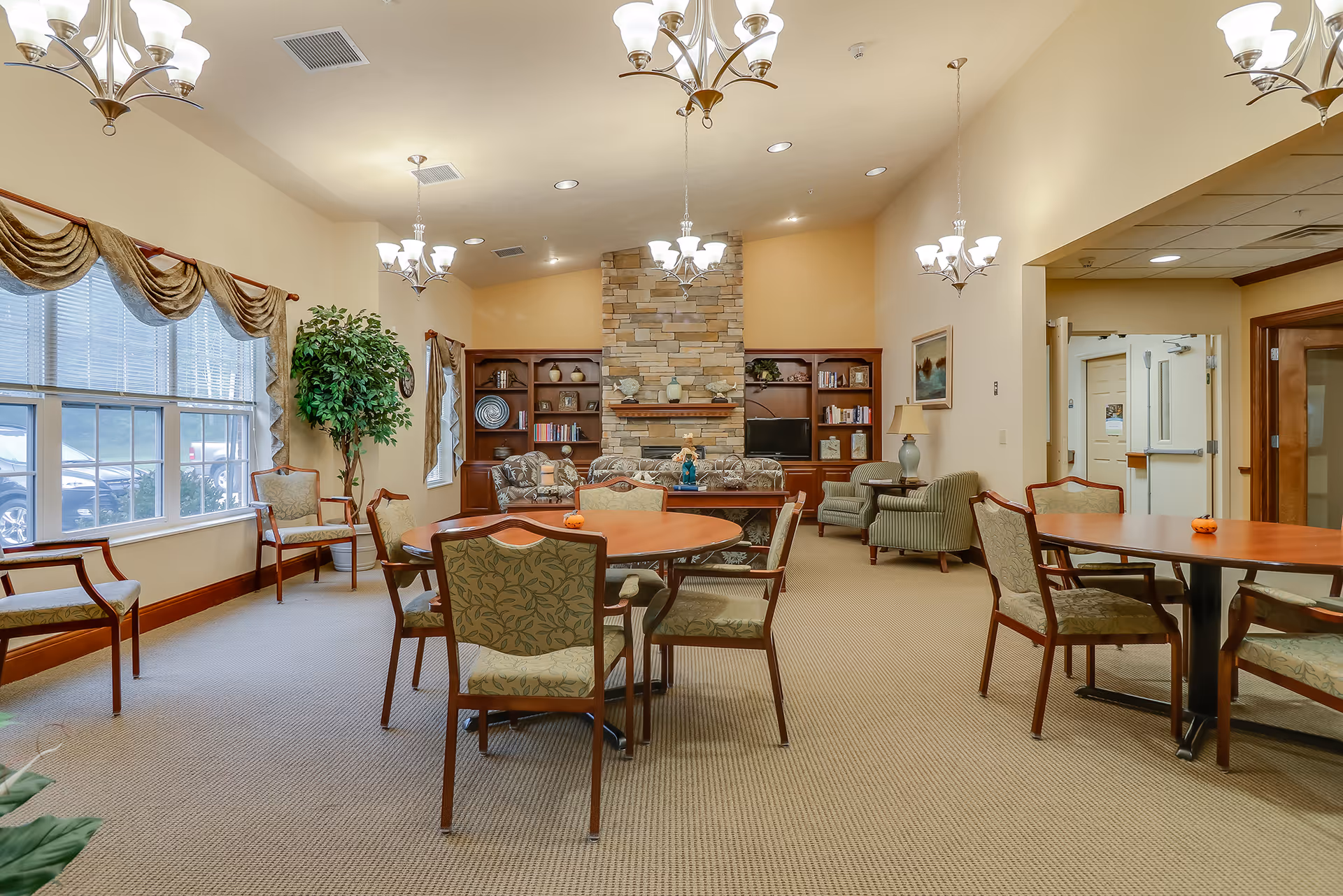 Well-lit assisted living common room with round tables and chairs, a stone fireplace, bookshelves, and large windows.