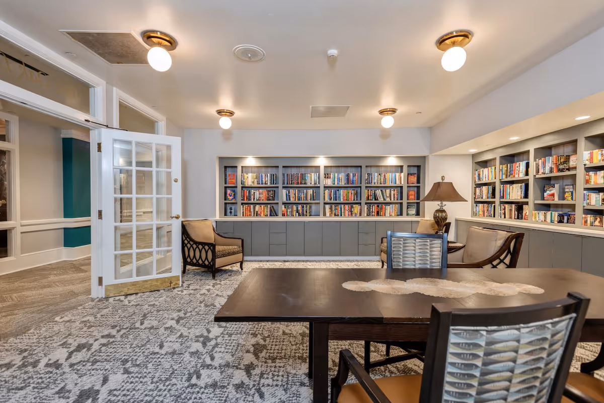 A cozy library room with built-in bookshelves filled with books along the back and right walls. There are two armchairs and a table with chairs in the foreground. The room has soft lighting from ceiling fixtures and a table lamp, with a patterned carpet covering the floor. A glass-paneled door is open to the left, leading to a hallway.