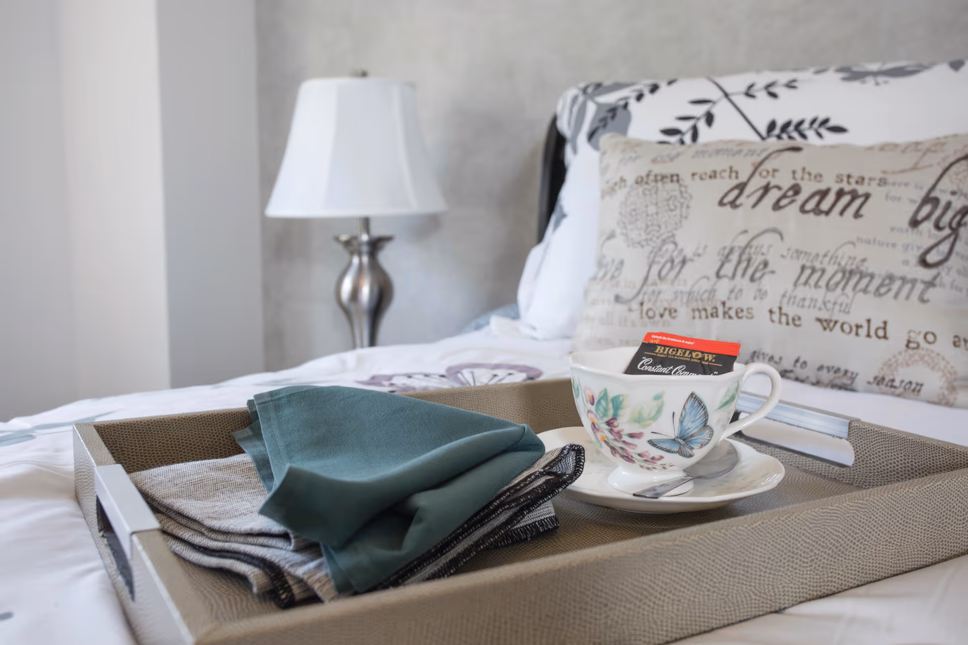 A decorative tray on a bed holding a floral teacup and folded napkins with a lamp and patterned pillow in the background.