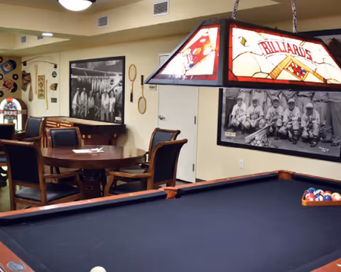 A recreational room featuring a pool table with billiard balls arranged on it, a round wooden table with chairs, vintage sports-themed black and white photographs on the walls, a jukebox, and a hanging stained glass billiards lamp.