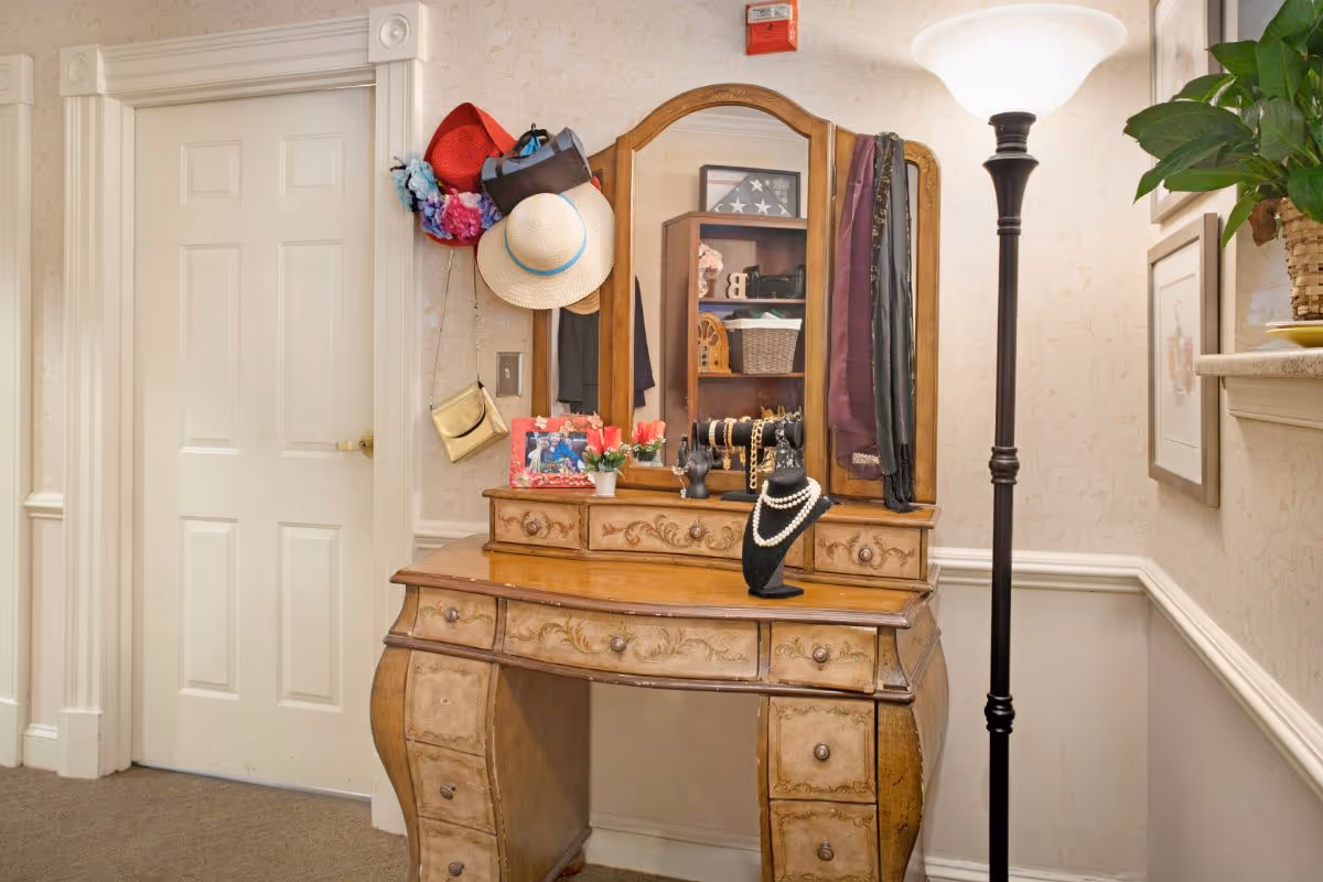 Ornate wooden vanity with a tri-fold mirror displaying hats, jewelry, and accessories in a wallpapered hallway beside a floor lamp.