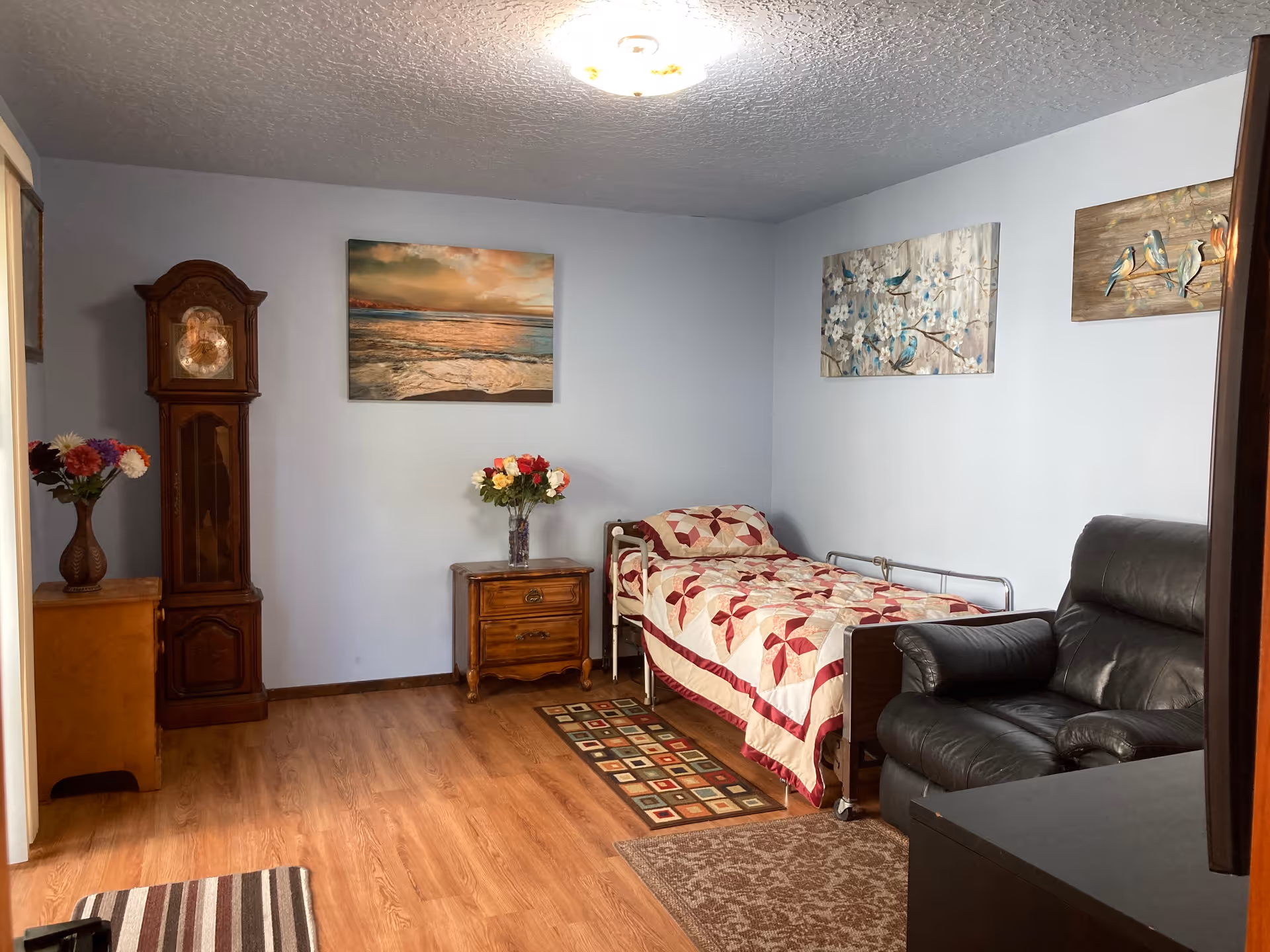 A cozy bedroom with a single bed covered by a quilt, a nightstand with flowers, a recliner, a grandfather clock, and wall art.