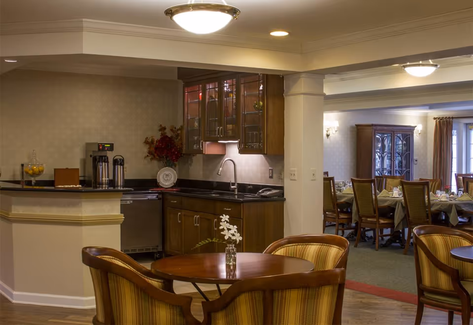 A communal dining area with round wooden tables and upholstered chairs, a small kitchenette with sink and cabinets, and a larger dining room visible in the background.