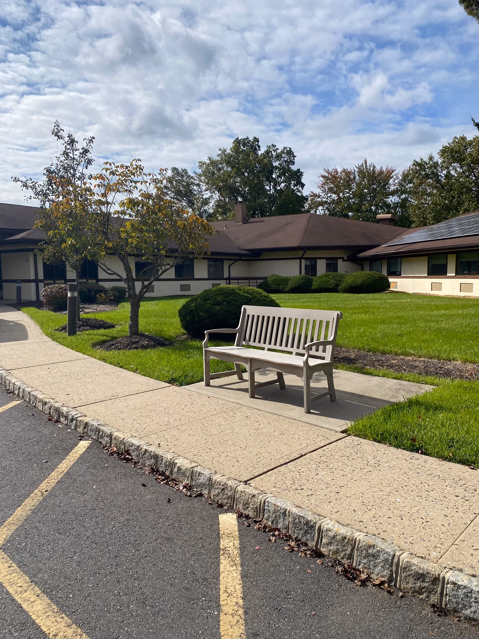 A bench on a sidewalk in front of a single-story senior living building with a manicured lawn, trees, and a partly cloudy sky.