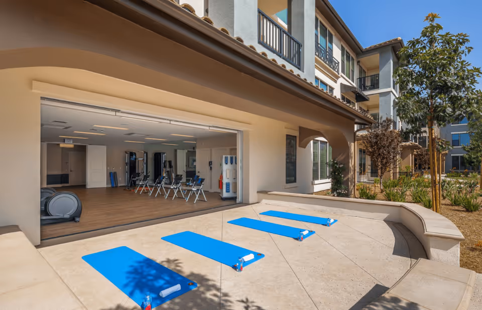 Outdoor patio area with four blue exercise mats laid out on the ground, each with a towel and water bottle. The patio is adjacent to a building with large open sliding doors revealing an indoor fitness room with exercise equipment and chairs. The building has multiple stories with balconies and windows, and there are trees and landscaping around the patio.