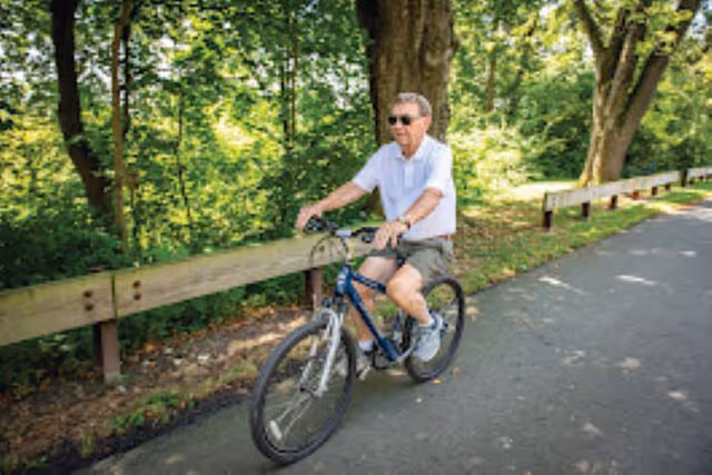 An elderly man wearing sunglasses, a white polo shirt, and shorts is riding a bicycle on a paved path surrounded by green trees and wooden guardrails.
