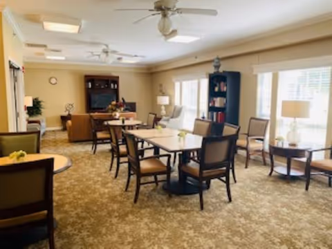 Bright communal dining and lounge area with tables, chairs, bookshelves and large windows.