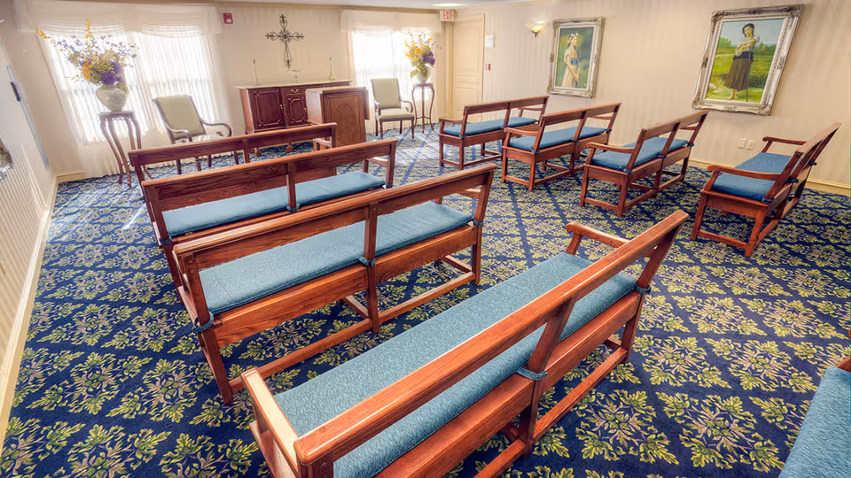 Carpeted chapel-style room with rows of wooden benches with blue cushions, paintings on the walls, and a lectern at the front.