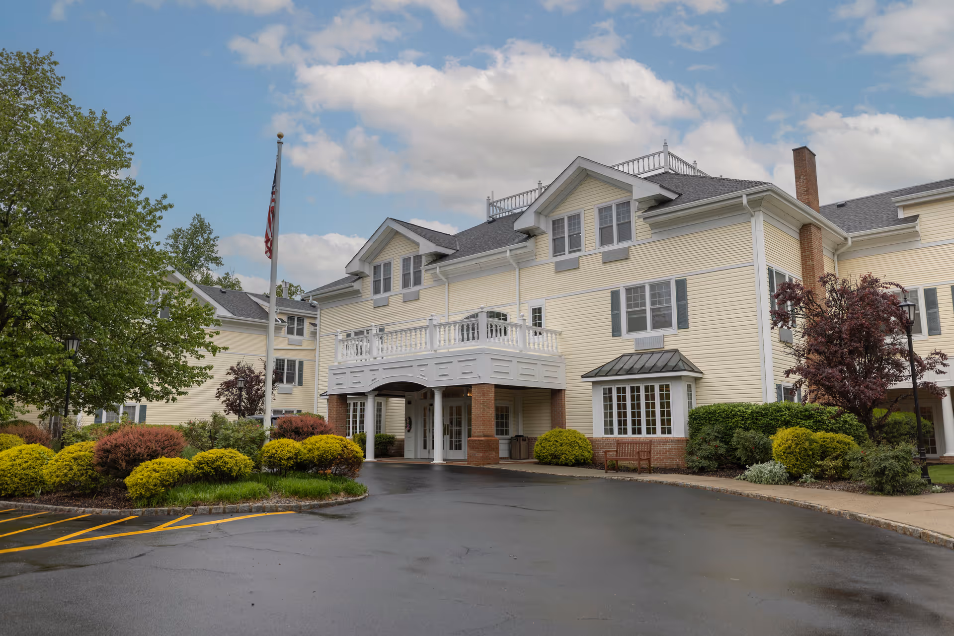 Exterior view of a large, light yellow assisted living facility building with white trim and multiple windows. The building has a covered entrance supported by brick columns and a balcony above it. There are well-maintained bushes and trees around the driveway, and an American flag is visible on a flagpole near the entrance. The sky is partly cloudy.