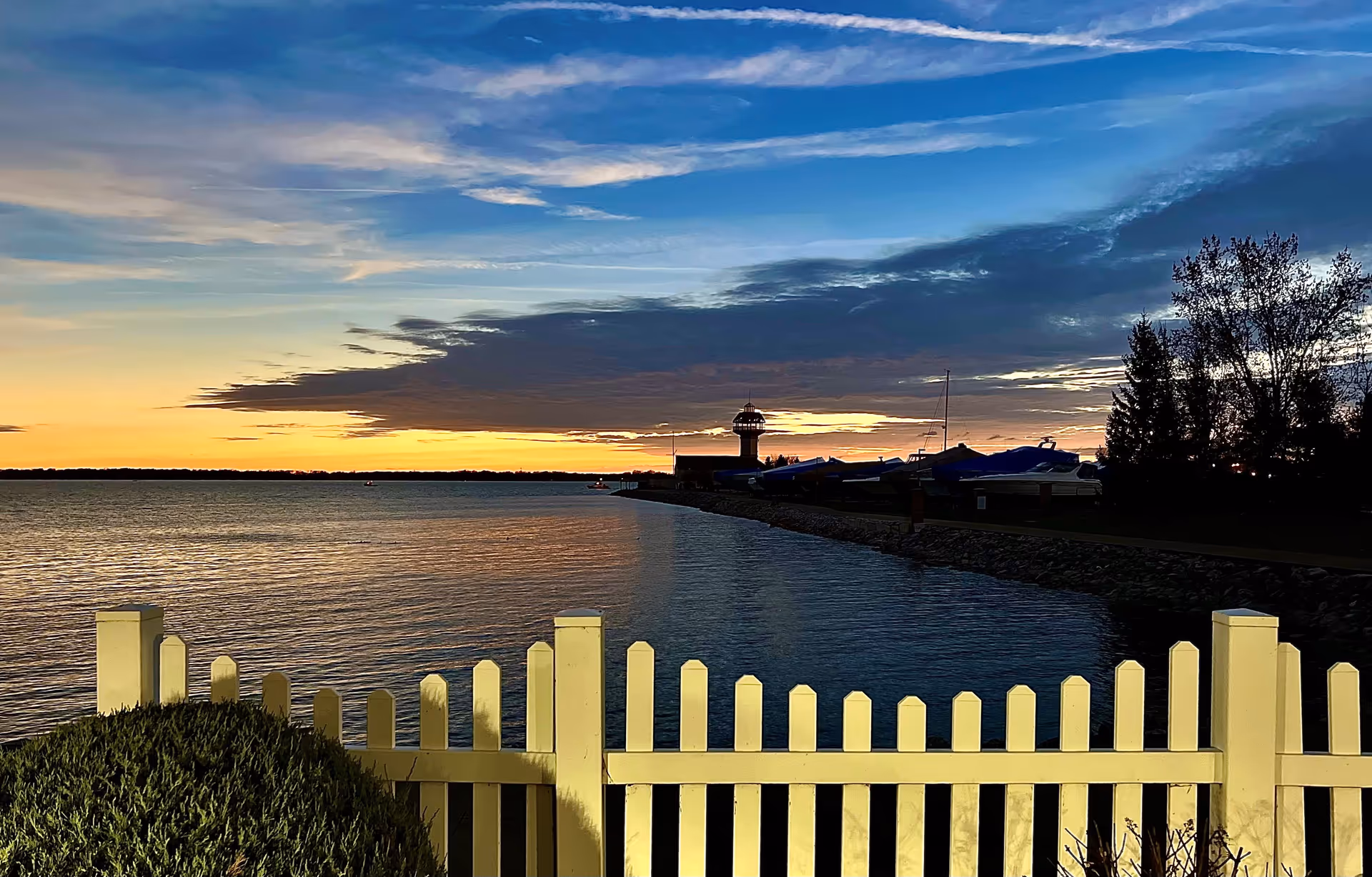 View of a waterfront at sunset with a yellow picket fence in the foreground, calm water reflecting the sky, a silhouette of a lighthouse and boats covered with tarps along the shoreline, and trees on the right side.