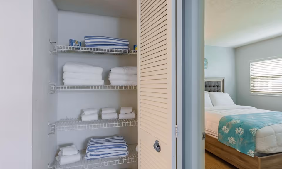 Open closet with white wire shelves holding neatly folded white and blue striped towels. To the right, a bedroom is visible with a bed made with white bedding and a blue blanket with a coral pattern, next to a window with closed blinds.
