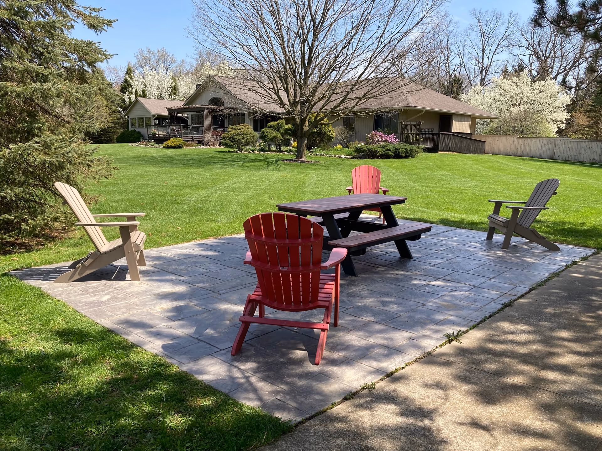 Outdoor patio area with a stone tiled floor featuring a dark wooden picnic table and four Adirondack chairs, two red and two beige. The patio is surrounded by a well-maintained green lawn, trees, and a single-story building in the background under a clear blue sky.