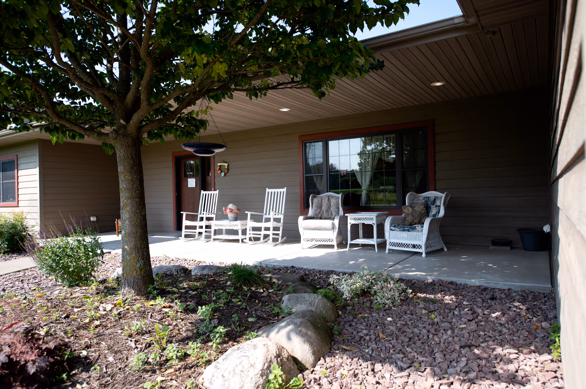 Covered front porch with white wicker and rocking chairs outside a single-story senior living building, with a tree and rock garden in front.