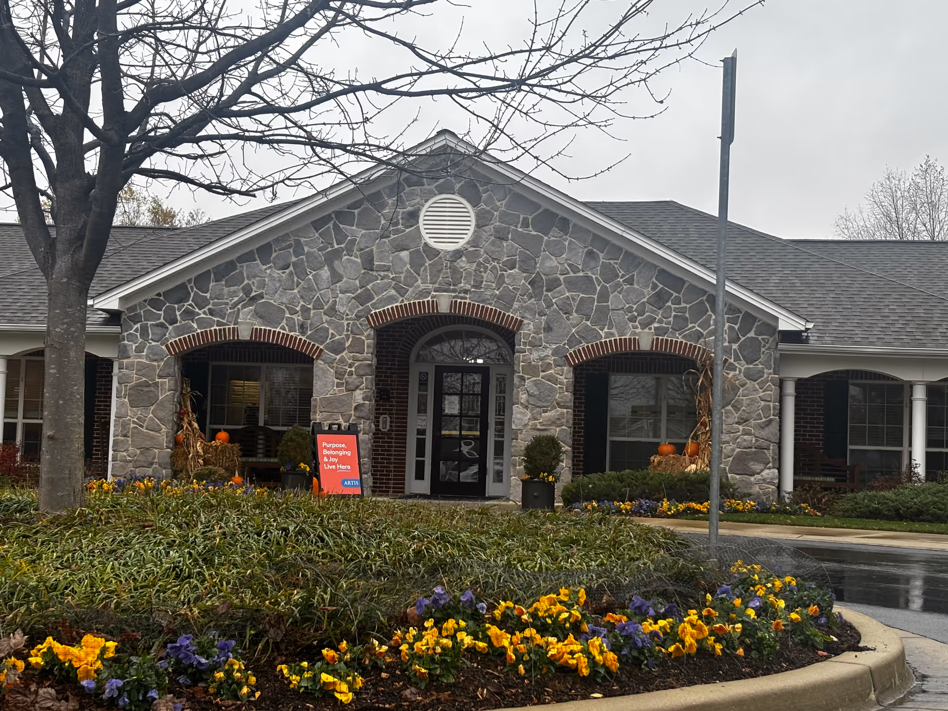Front exterior view of a stone-faced building with a peaked roof and arched doorways. There are flower beds with yellow and purple flowers in front, a tree without leaves, and a sign near the entrance that reads 'Purpose, Belonging & Joy Live Here ARTIS'. The sky is overcast.