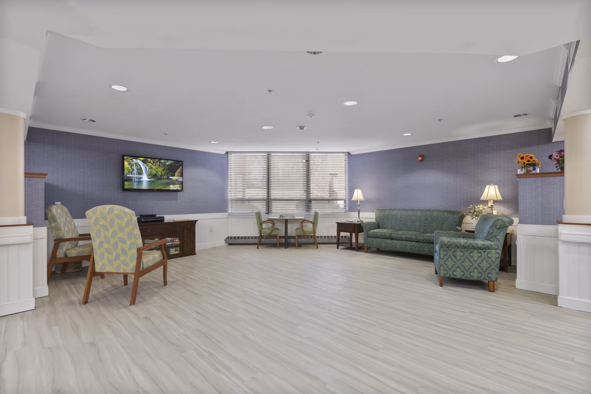 A spacious and well-lit common area in a senior living facility with light wood flooring, purple patterned walls, and white wainscoting. The room features a green patterned sofa and armchair, two yellow and green patterned chairs, a small table with two green chairs near a window with blinds, two side tables with lamps, a flat-screen TV mounted on the wall displaying a waterfall scene, and decorative flowers on shelves.