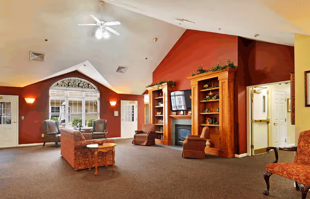 A spacious senior living facility common area with high vaulted ceilings and warm lighting. The room features a red accent wall with two wooden bookshelves flanking a fireplace and a mounted TV. There are several upholstered chairs and a sofa arranged around a small wooden table. Large windows and doors allow natural light to enter the room.