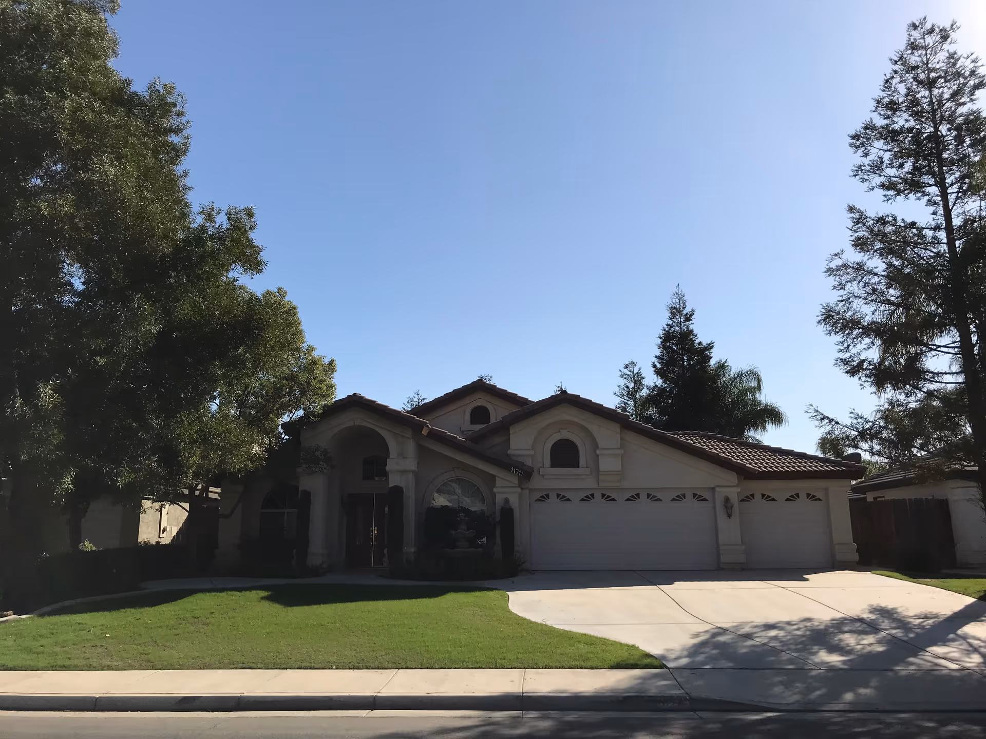 Front exterior view of a single-story residential-style building with a three-car garage, arched windows, and a tiled roof, surrounded by trees and a well-maintained lawn under a clear blue sky.
