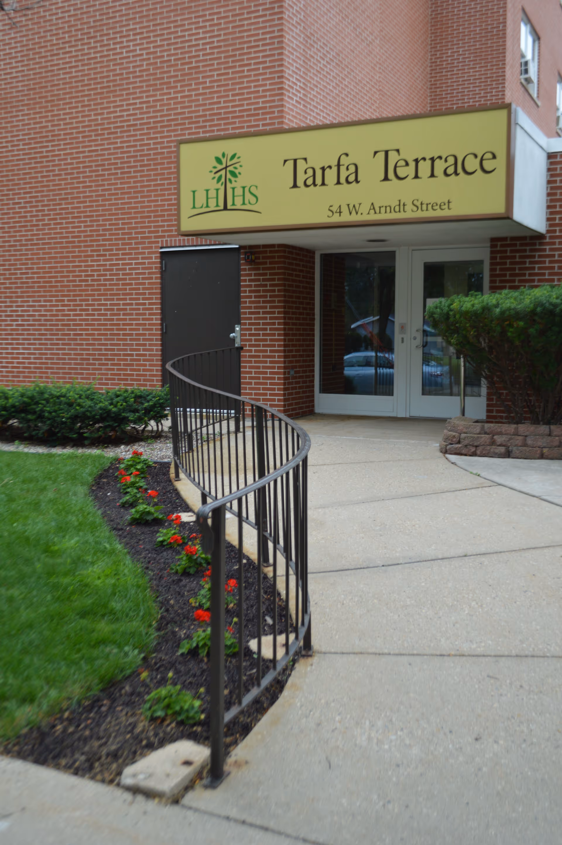 Entrance to Tarfa Terrace Assisted Living facility with a yellow sign above the door displaying the name and address 54 W. Arndt Street. The entrance has double glass doors, a curved black metal railing, and landscaping with green grass and red flowers.