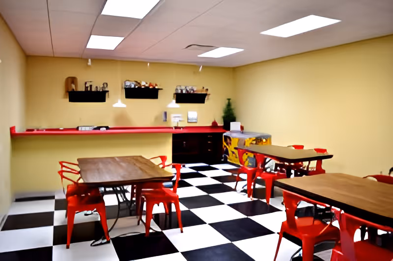 A dining area with black and white checkered floor tiles, yellow walls, and a ceiling with fluorescent lights. There are several wooden tables surrounded by red metal chairs. A red counter runs along one wall with shelves above it holding decorative items. A small Christmas tree and a pinball machine are visible in the corner.