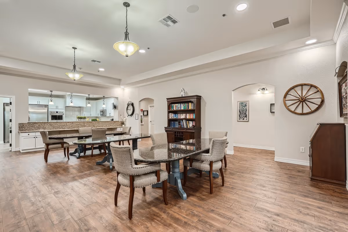A spacious, well-lit common area with a dining table and chairs in the foreground, a kitchen with white cabinets and granite countertops in the background, wooden flooring, and decorative elements including a bookshelf, wall clock, and a wooden wagon wheel mounted on the wall.