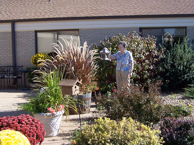 An elderly woman stands in a sunny garden courtyard outside a Wheatlands Health Care Center, surrounded by potted plants, flowers, and a birdhouse nearby.