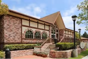 Front exterior of a brick senior care building with Tudor-style trim, arched windows, entry stairs, landscaping, and lampposts under a blue sky.