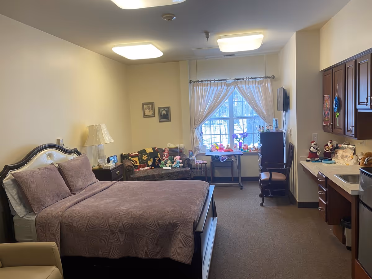 A cozy bedroom in a senior living facility with a large bed covered in a purple quilt and matching pillows. There is a nightstand with a lamp beside the bed, a small couch with stuffed animals, a window with white curtains letting in natural light, a small table with more stuffed animals, a chair, and a kitchenette area with cabinets, a sink, and a mini fridge.