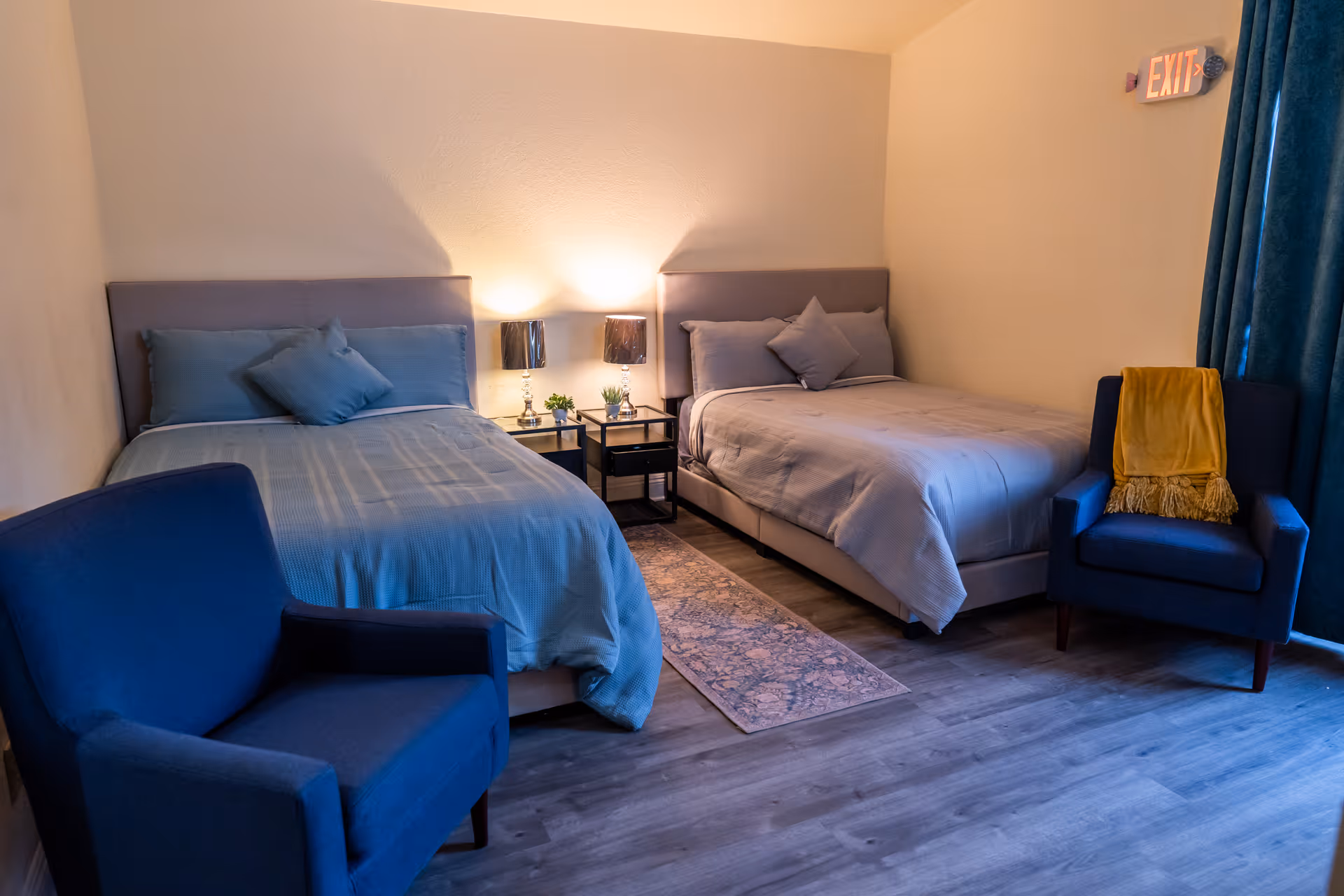 Two neatly made twin beds in a softly lit bedroom with a nightstand and lamps between them and two blue armchairs.