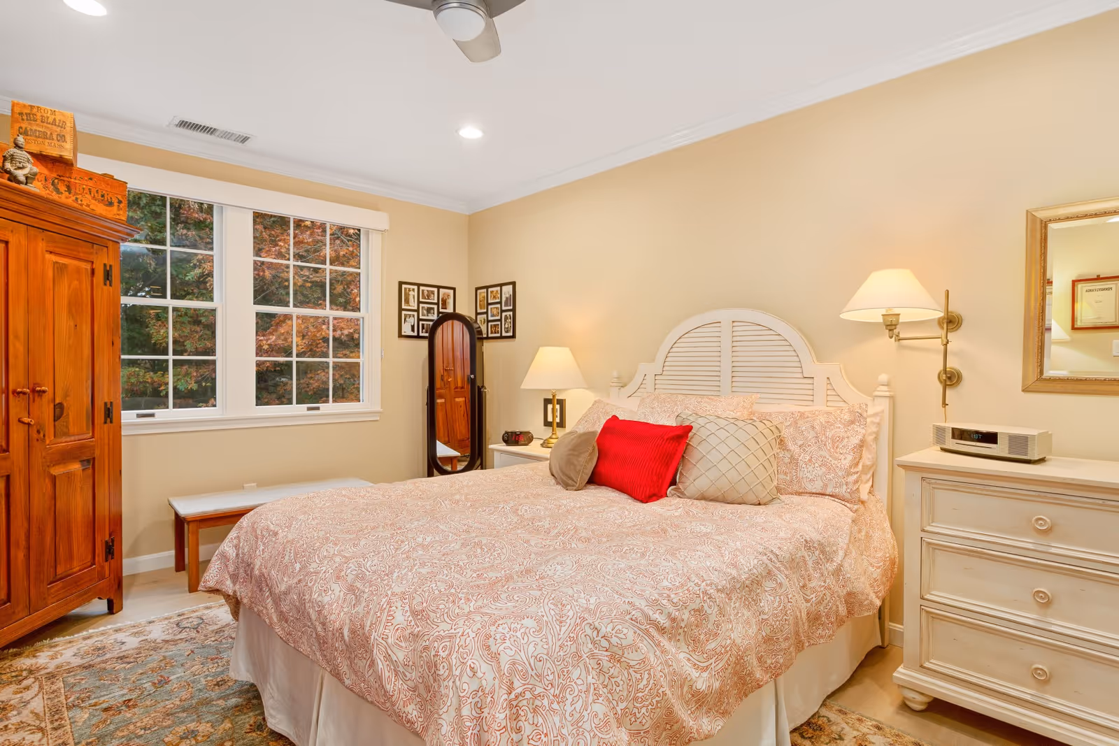 A cozy bedroom with a large bed covered in a patterned bedspread and multiple pillows, including a red one. There is a wooden armoire on the left side, a bench under a window with white frames showing autumn foliage outside, and a standing mirror in the corner. On the right side, there is a white dresser with a lamp and a mirror above it. The walls are painted a soft beige color.