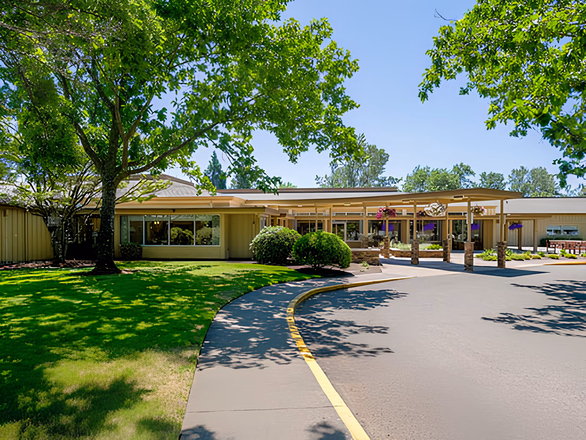 Exterior view of Avamere Riverpark of Eugene facility showing a single-story building with a covered entrance, surrounded by green trees and a well-maintained lawn under a clear blue sky.