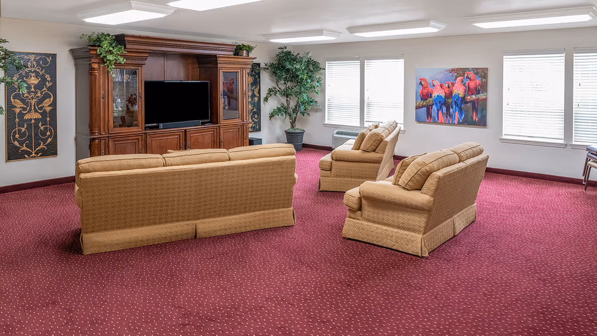 Spacious common room with beige sofas and armchairs arranged toward a wooden entertainment center and TV, red patterned carpet and windows along one wall.