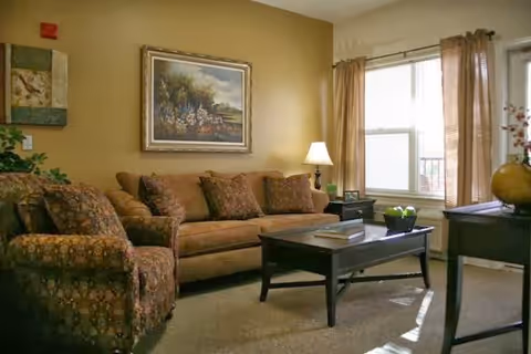 Sunlit living room with a sofa, patterned armchair, coffee table, table lamp, and framed painting on the wall.